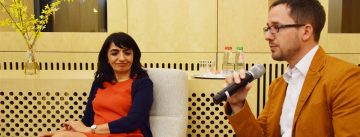 Kerim Arpad vom DTF spricht in ein Mikrofon am rechten Bildrand während eine Frau am linken Bildrand in einem roten Kleid verlegen in seine Richtung lächelt